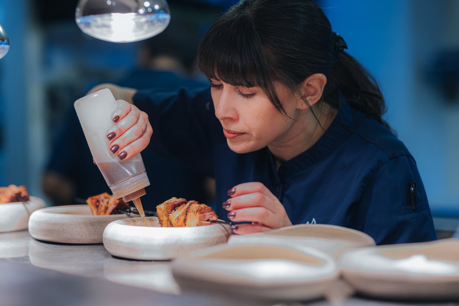 Elena finishing a plate, dressing the dish under the pass lamp